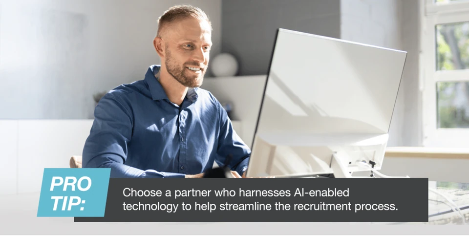 Smiling man in a blue shirt works on a computer by a sunny window. Text overlay: "PRO TIP: Choose a partner for AI recruitment tech."