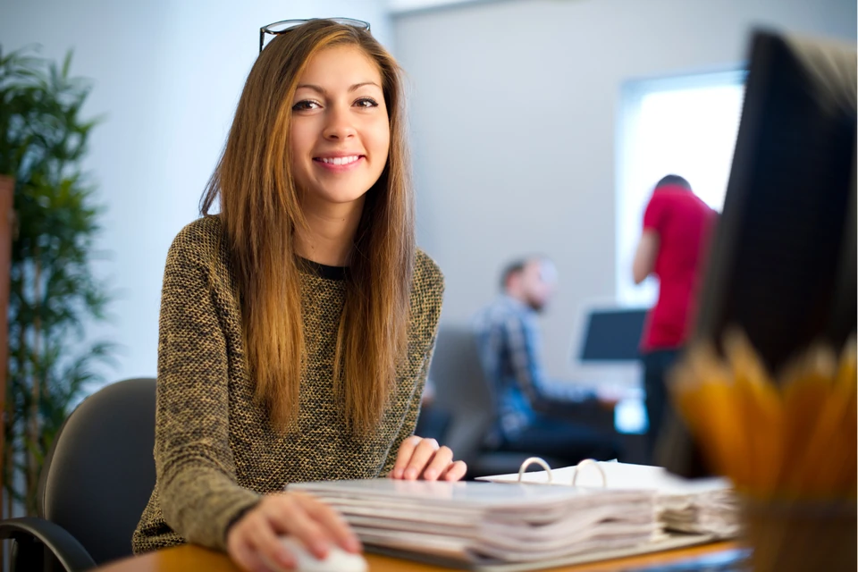 Smiling young woman at an office desk, with blurred coworkers and a computer in the background, in a bright workspace.